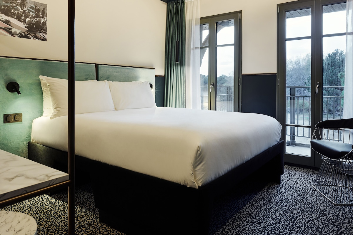 Habitación de hotel moderna con una cama grande, un escritorio y una silla. Se ve una ventana con vistas al exterior.