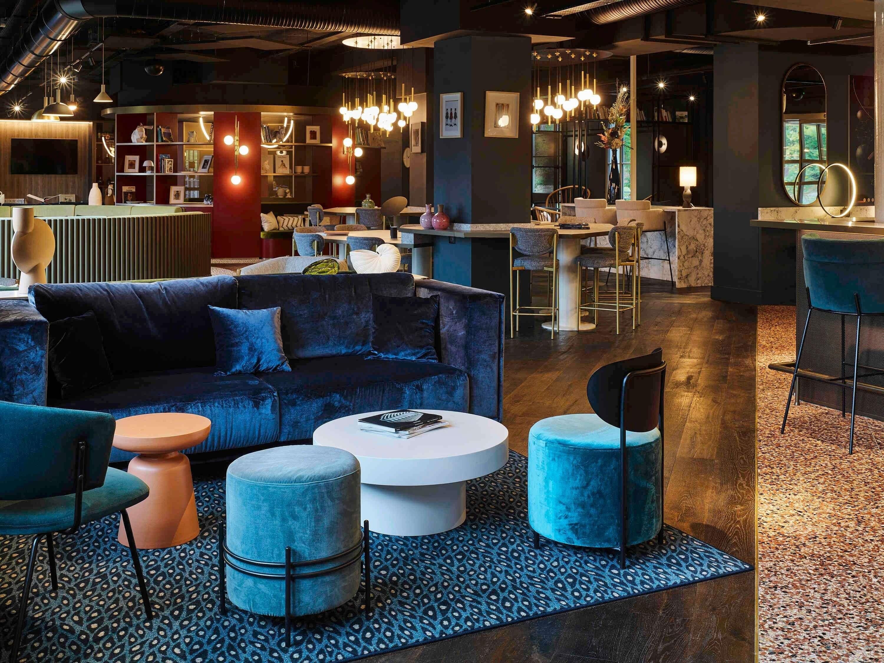 A modern lounge area with a dark blue sofa, round ottomans, and a white coffee table. A bar area with stools and a mirror is visible in the background.