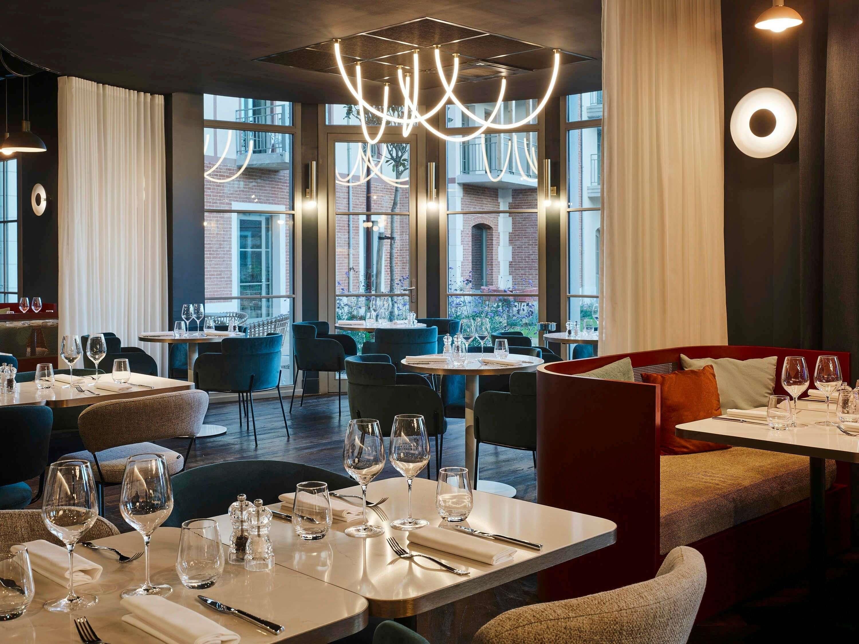 L’intérieur d’un restaurant, avec des tables dressées pour le service, de grandes fenêtres et des luminaires modernes.