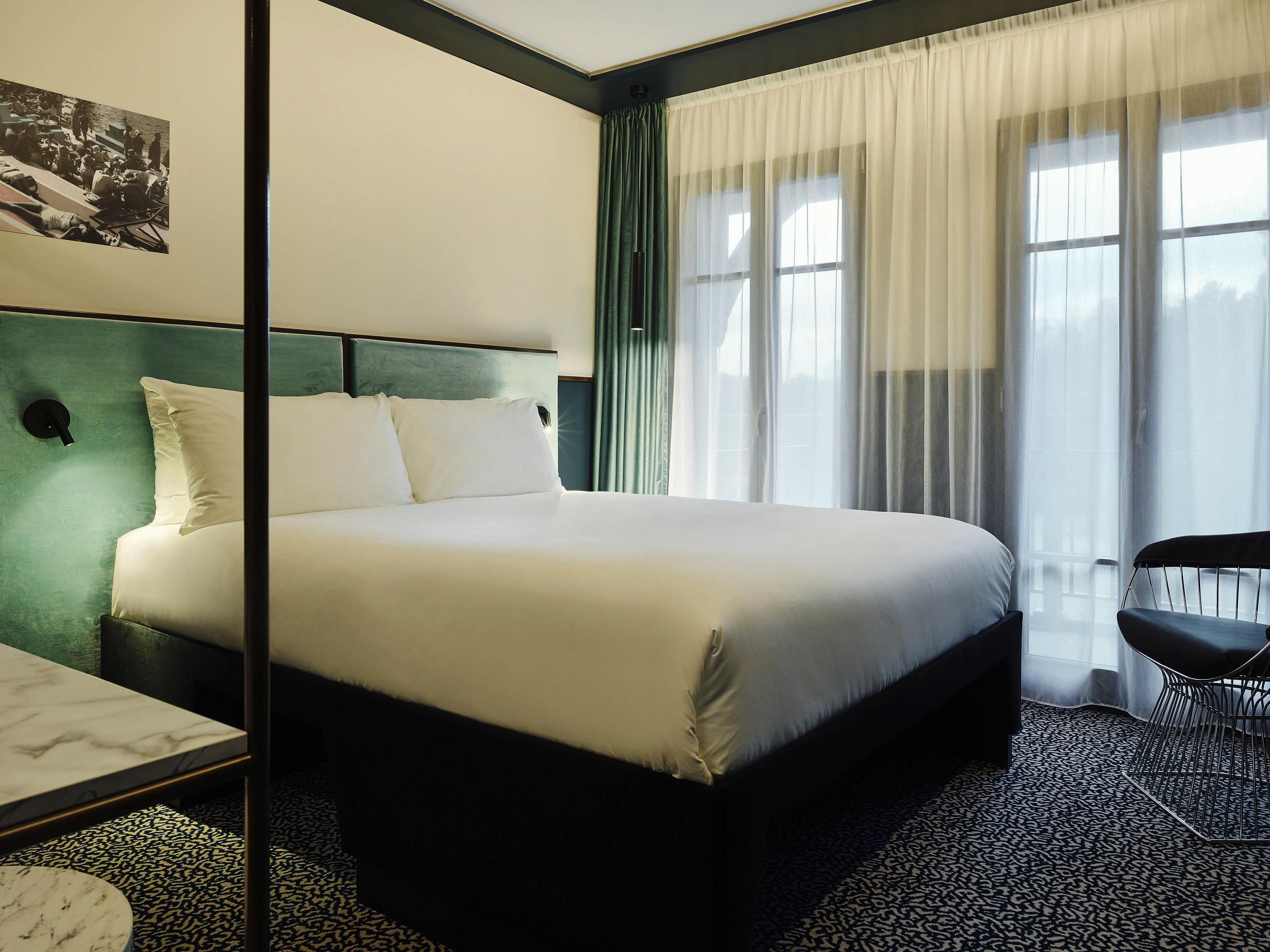 A hotel room with a bed, a desk, a chair, a window with curtains, and a wall-mounted light.