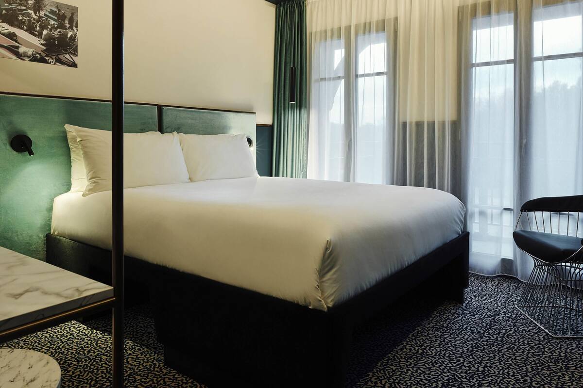 Habitación de hotel con cama, escritorio, silla, ventana con cortinas y una lámpara en la pared.
