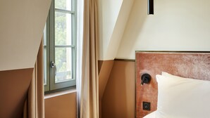 Habitación de hotel con cama, una mesita redonda, ventana con cortinas y panel de control en la pared.