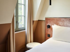 Habitación de hotel con cama, una mesita redonda, ventana con cortinas y panel de control en la pared.