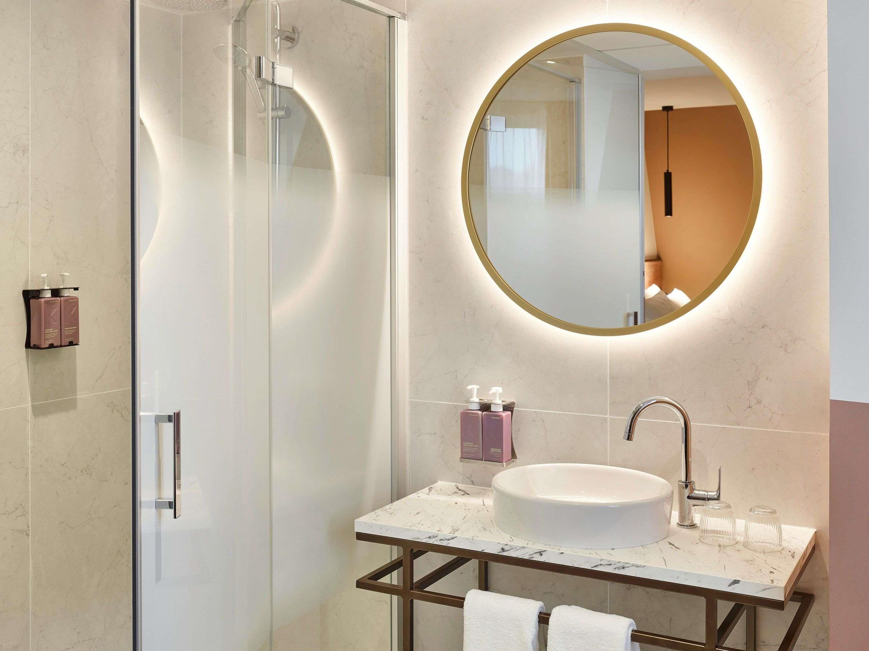 Un baño moderno con cabina de ducha de vidrio, espejo redondo, lavabo de mármol y toallero.