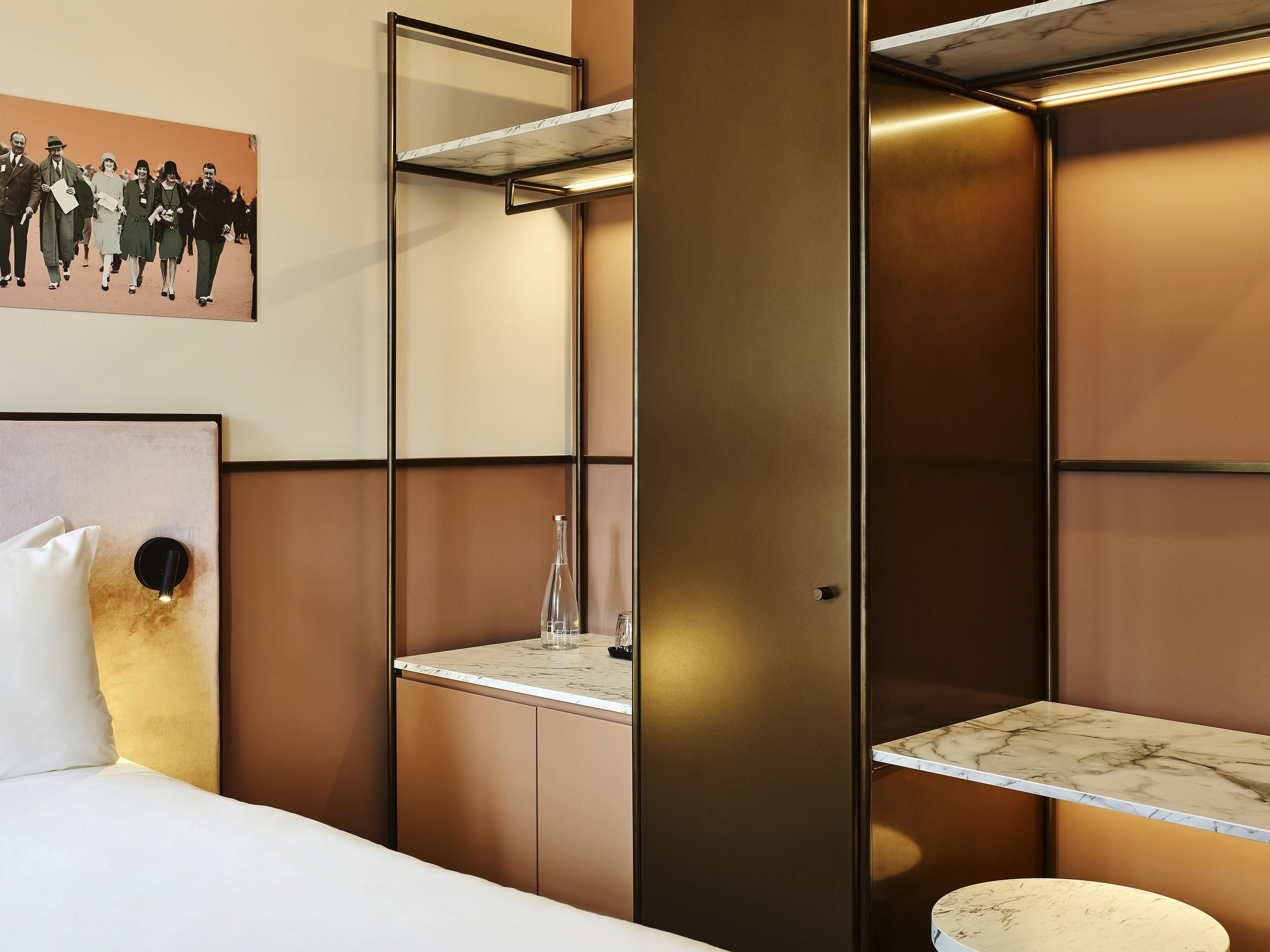 A hotel room with a bed, a nightstand, a marble-topped table, a shelf with a mirror, and a wall-mounted picture.