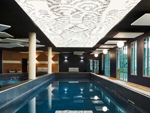 Una piscina interior con techo decorado con un mosaico, grandes ventanales a un lado y una zona de estar con sillas y un banco.