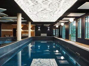 Una piscina interior con diseño en el techo, ventanas amplias y una zona de estar.