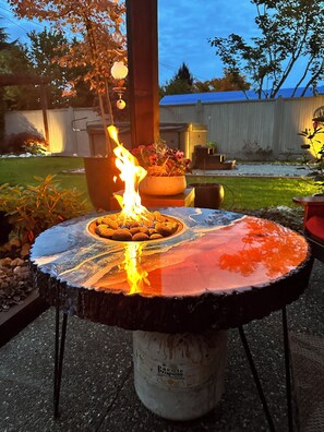 A round table with a glass top, a fire burning on it, and a potted plant with pink flowers in the background.