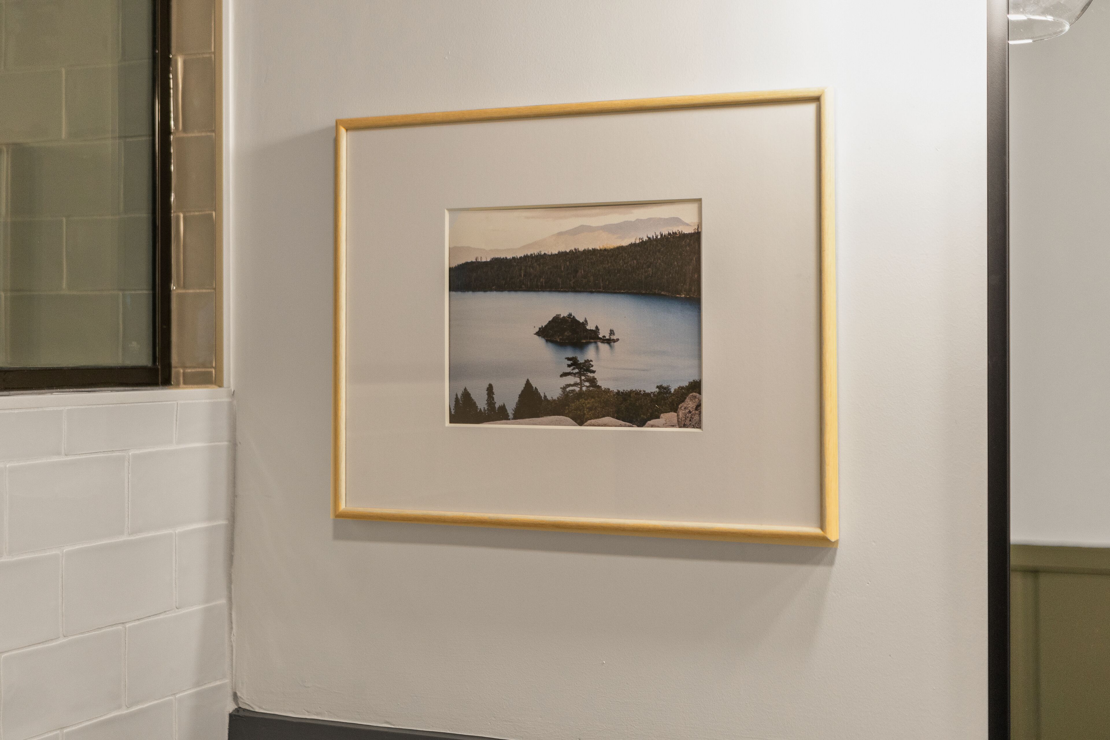 Pintura emoldurada de uma paisagem com um lago, uma pequena ilha e montanhas ao fundo, pendurada em uma parede branca.
