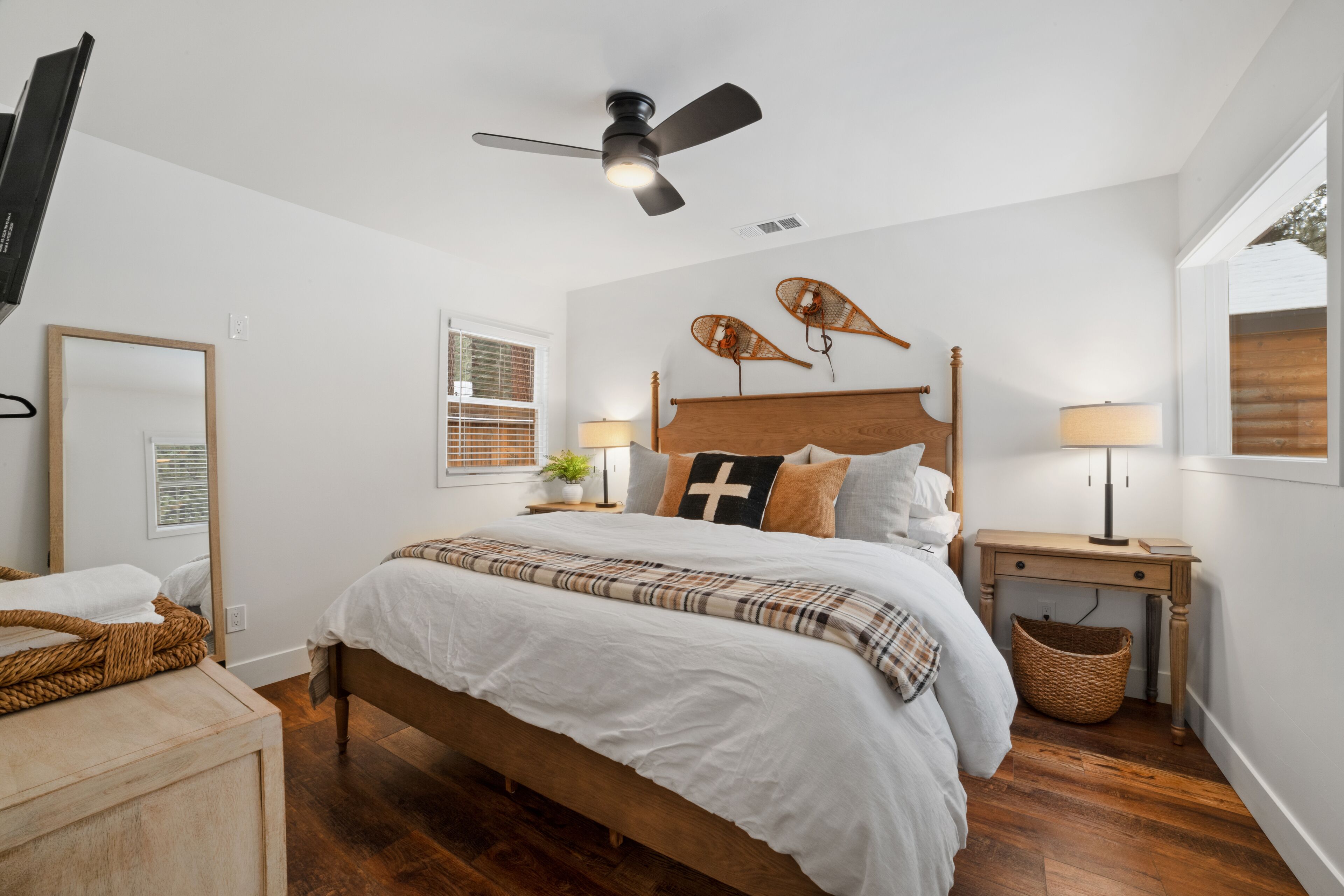 A bedroom with a wooden bed, white bedding, a ceiling fan, a bedside table with a lamp, a mirror, and a window with blinds.
