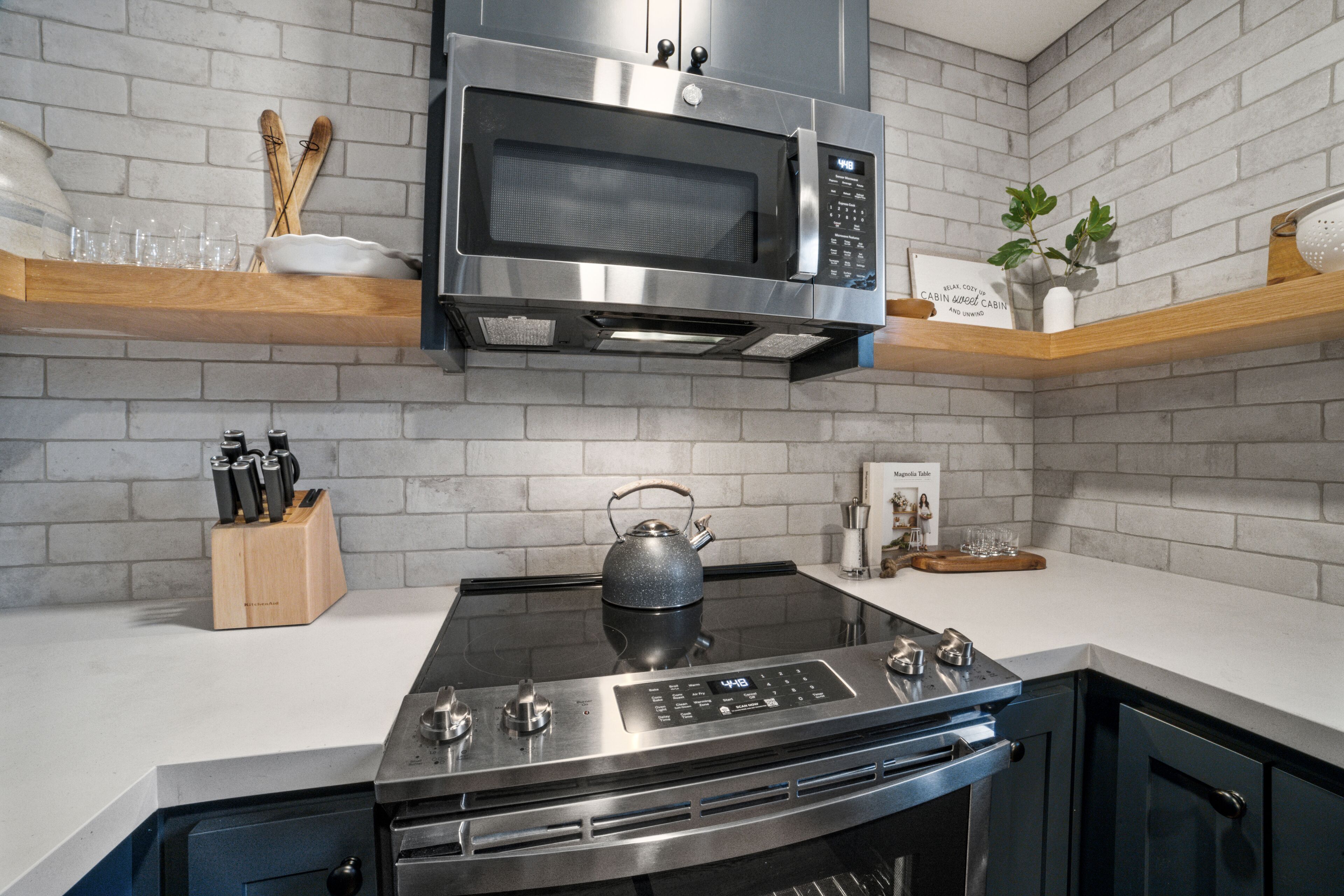 A kitchen with a stainless steel microwave, a gas stove, and a kettle. There are wooden shelves with glassware and a knife block.