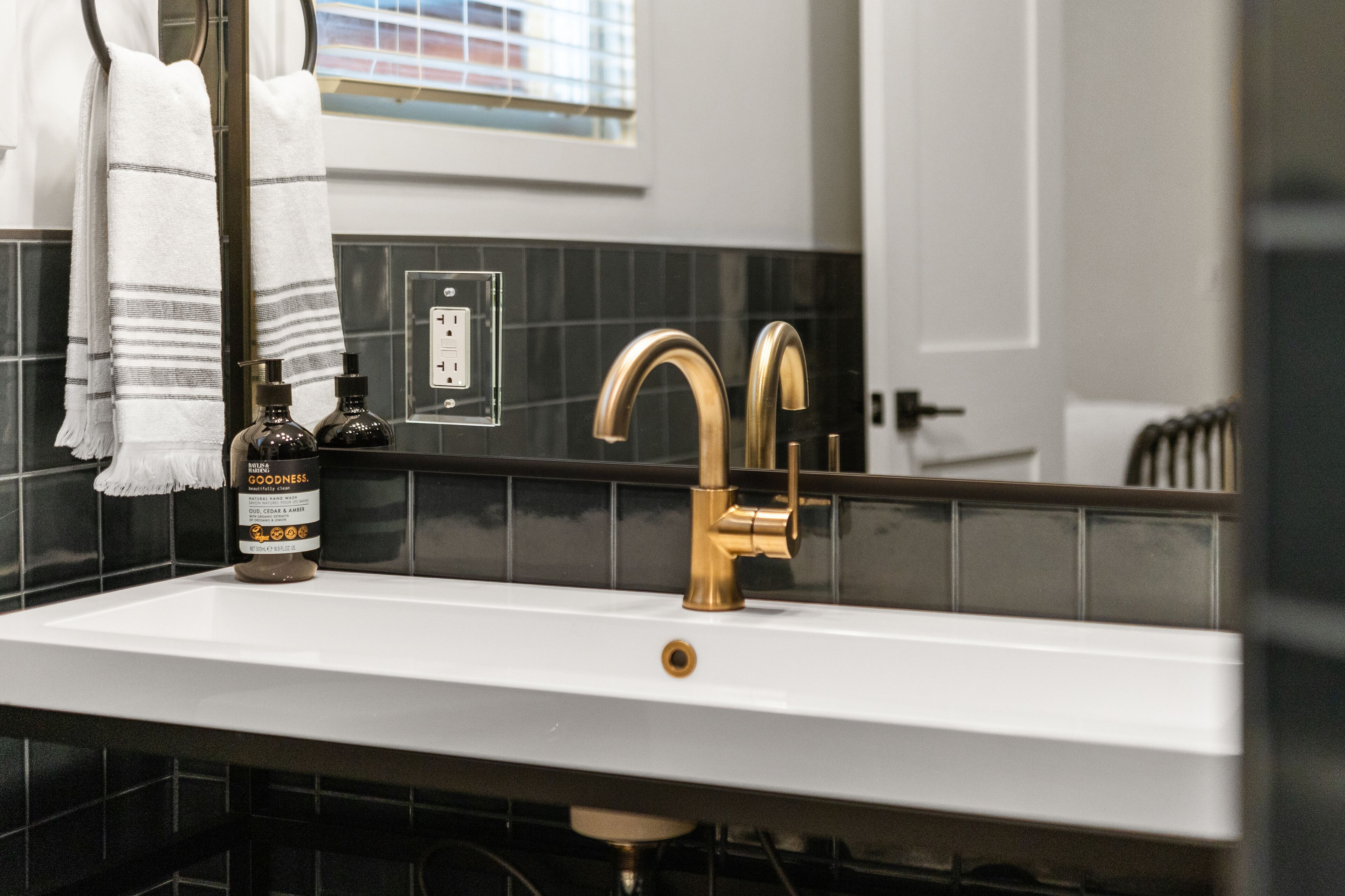 A bathroom with a white sink, gold faucet, and black tiled walls. There is a towel hanging on the wall and a bottle of hand soap on the counter.