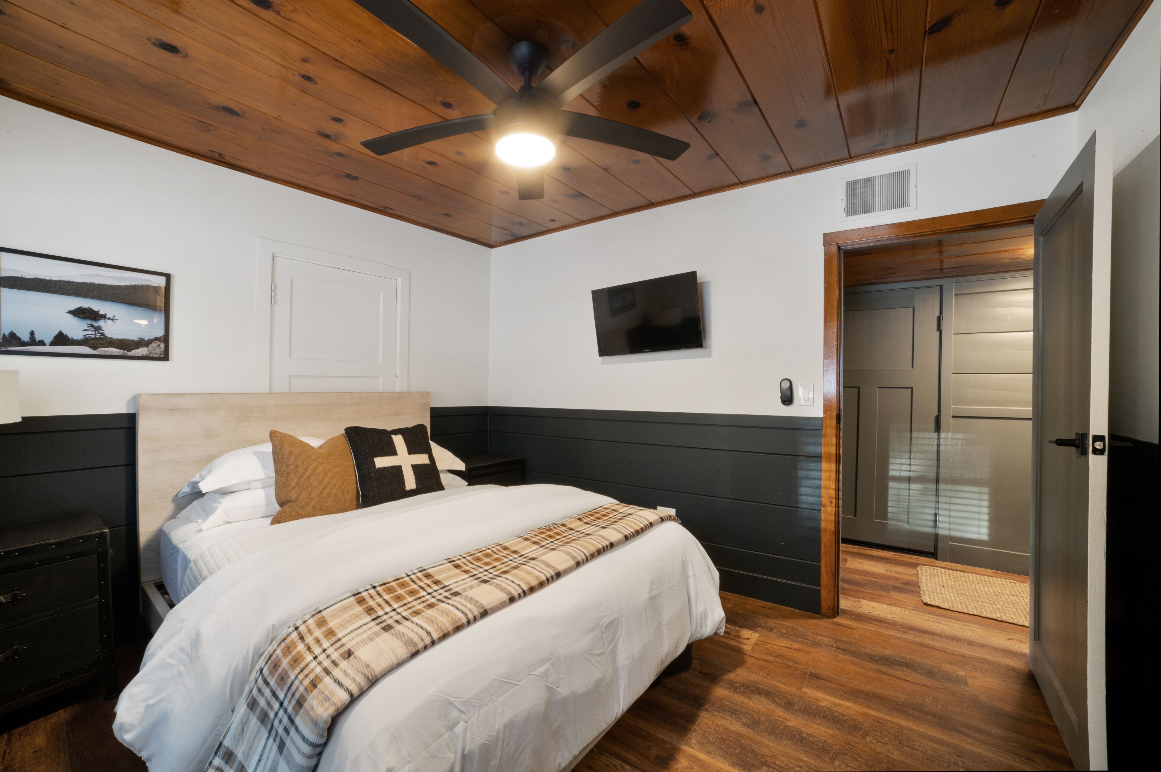 Un dormitorio con una cama, ventilador de techo, televisión, puerta y un cuadro en la pared.