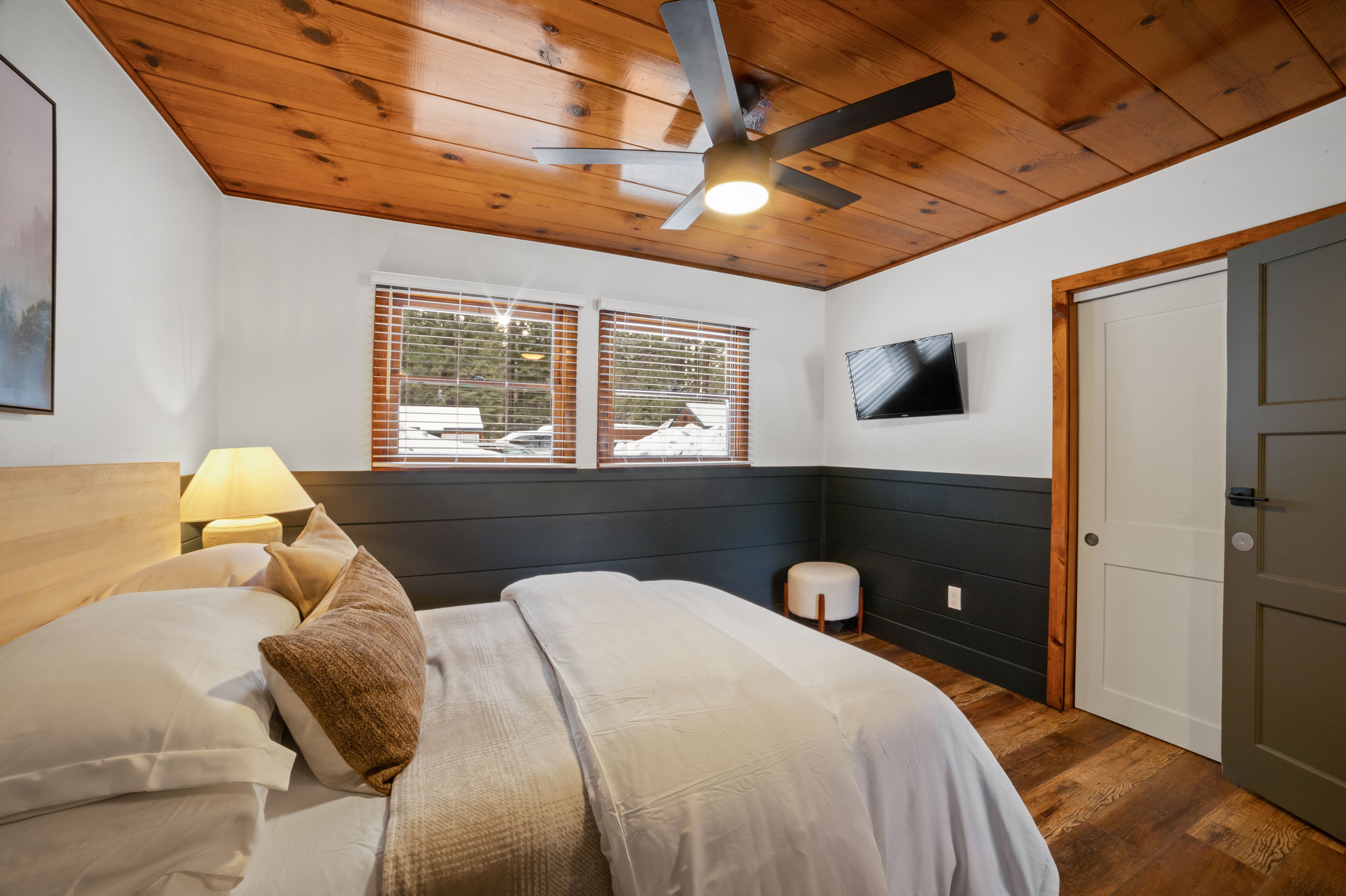 A bedroom with a wooden ceiling, a bed with white bedding, a nightstand with a lamp, a wall-mounted TV, and a door.
