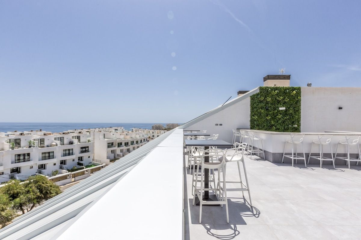 A rooftop terrace with outdoor seating, a view of the sea, and a modern building with a green wall.