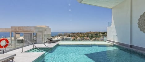 A rooftop infinity pool with clear blue water, a lifebuoy, and a view of a coastal town.
