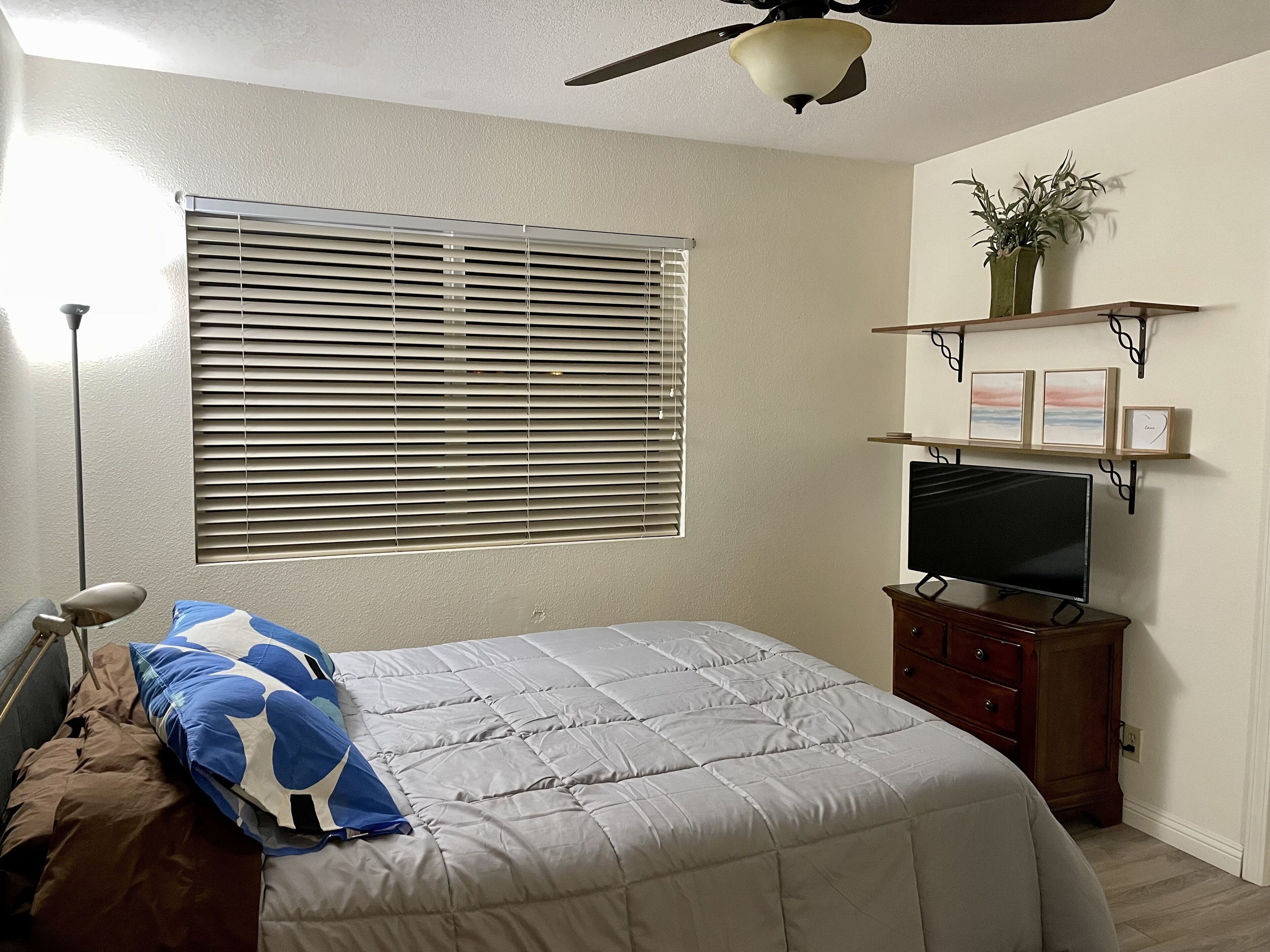 A bedroom with a bed, a ceiling fan, a window with blinds, a TV, a dresser, and a shelf with decorative items.