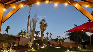 Un patio al aire libre con una pérgola de madera, luces colgantes, palmeras y un paraguas rojo.