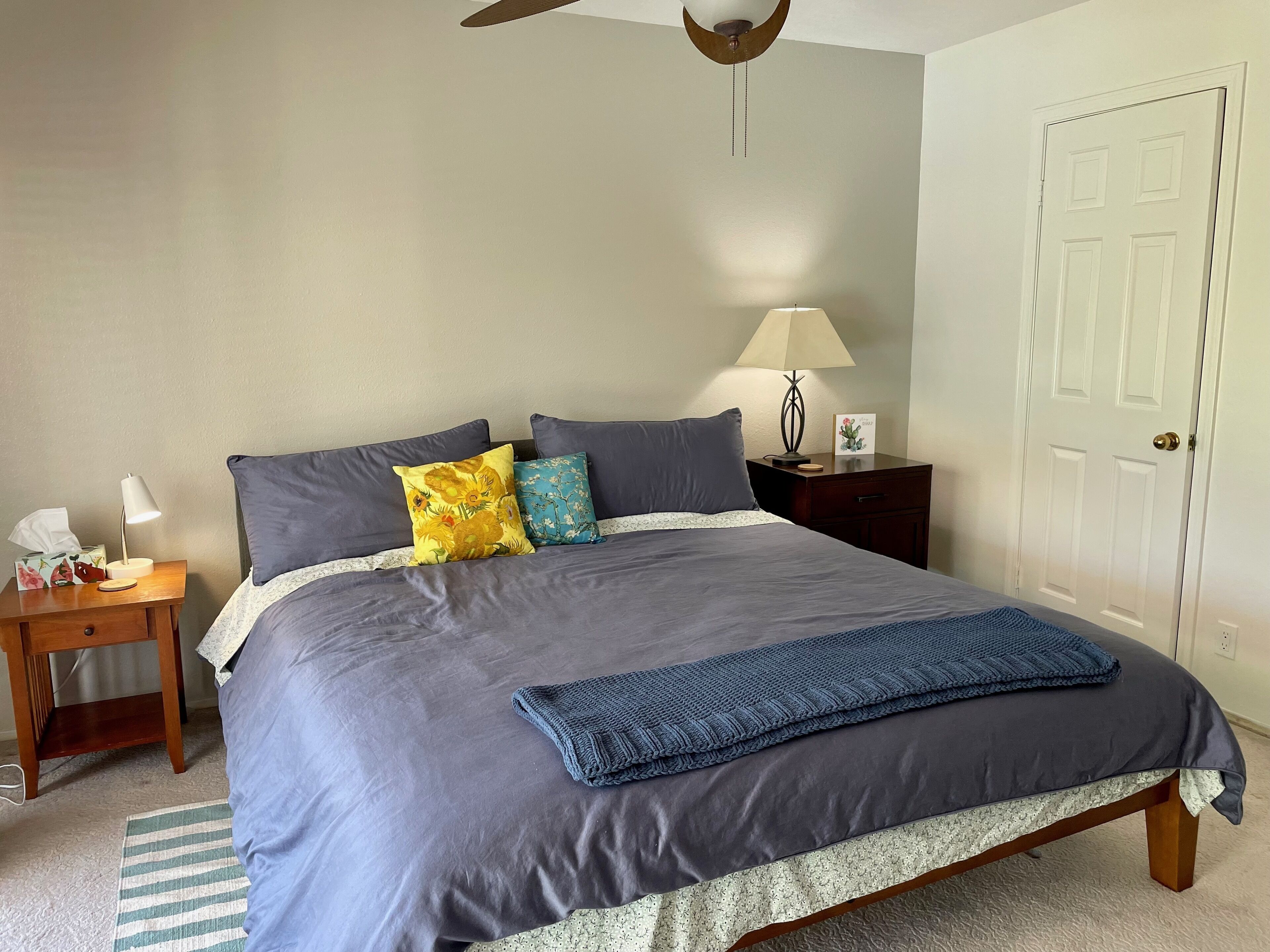 Un lit bien fait, recouvert d’une couvre-lit bleu foncé et agrémenté d’oreillers, une lampe de chevet et une petite table de nuit sur laquelle se trouvent une boîte de mouchoirs et une lampe.