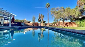 Una piscina cristalina que refleja el entorno circundante, incluyendo un edificio con terraza cubierta y asientos al aire libre.