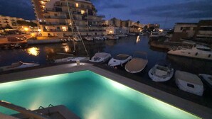 A marina at dusk with boats docked and a lit swimming pool in the foreground.
