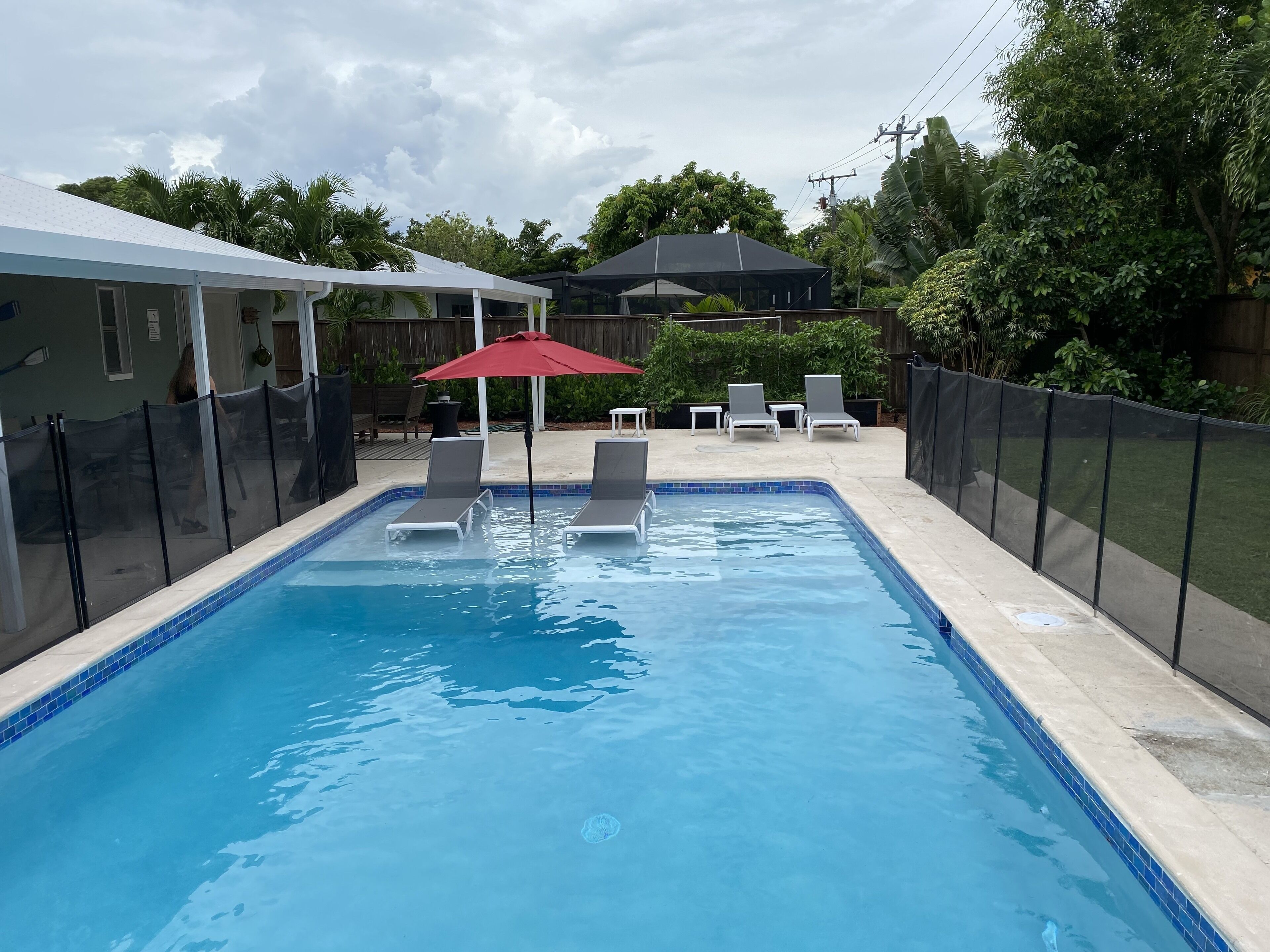 Une piscine rectangulaire aux bords recouverts de carreaux bleus, entourée d’une clôture noire. On y trouve deux chaises longues et un parasol rouge, placés sous un espace ombragé.