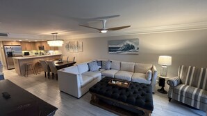 Een moderne woonkamer met een hoekbank, een zwart gestoffeerd pouf, een gestreept fauteuil en een eethoek, met een keuken op de achtergrond.