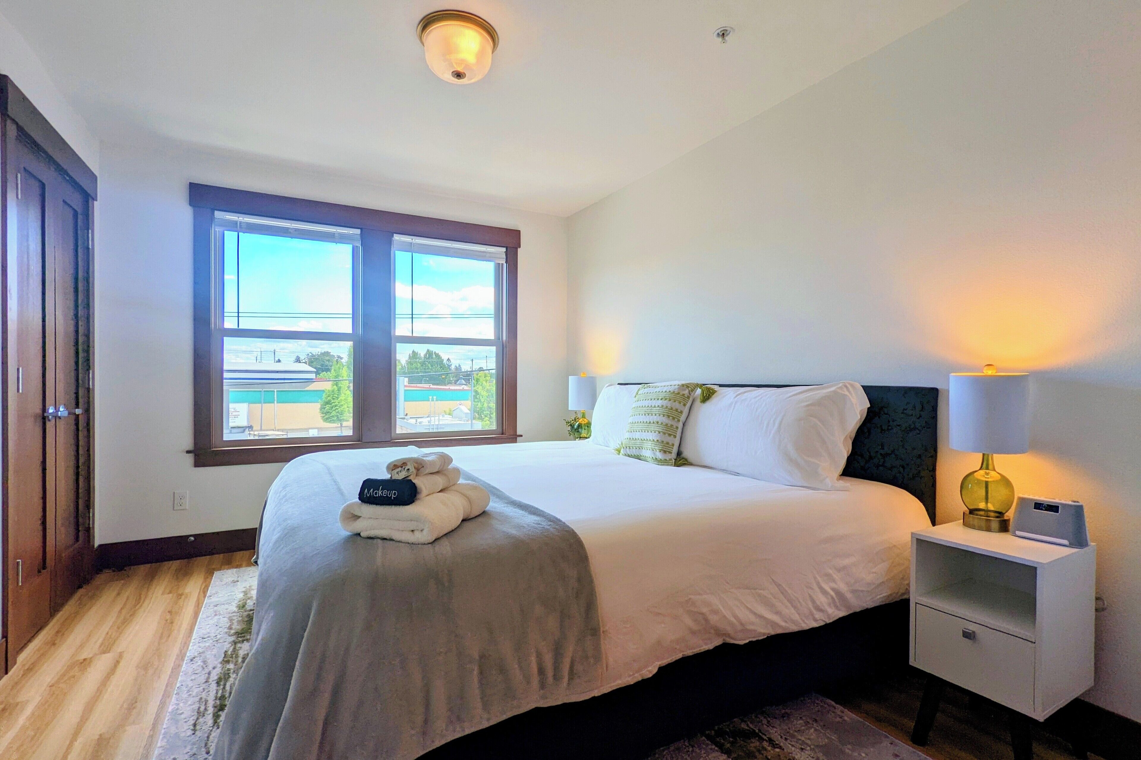 Una cama bien tendida con una toalla doblada y una almohada, una mesita de noche con una lámpara y una ventana con vistas al exterior.