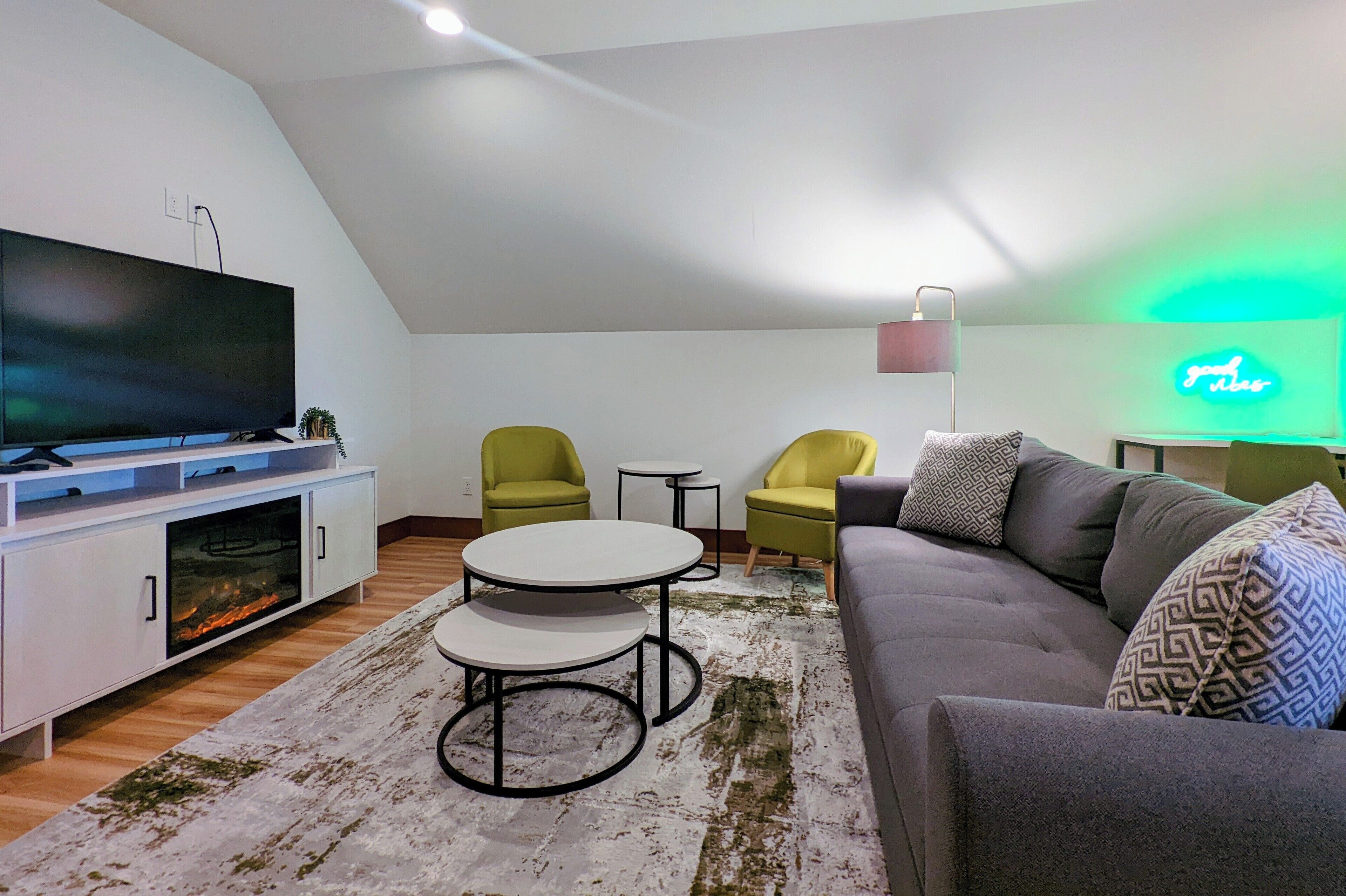 A modern living room with a flat-screen TV, two yellow chairs, a grey sofa, and a small round table.