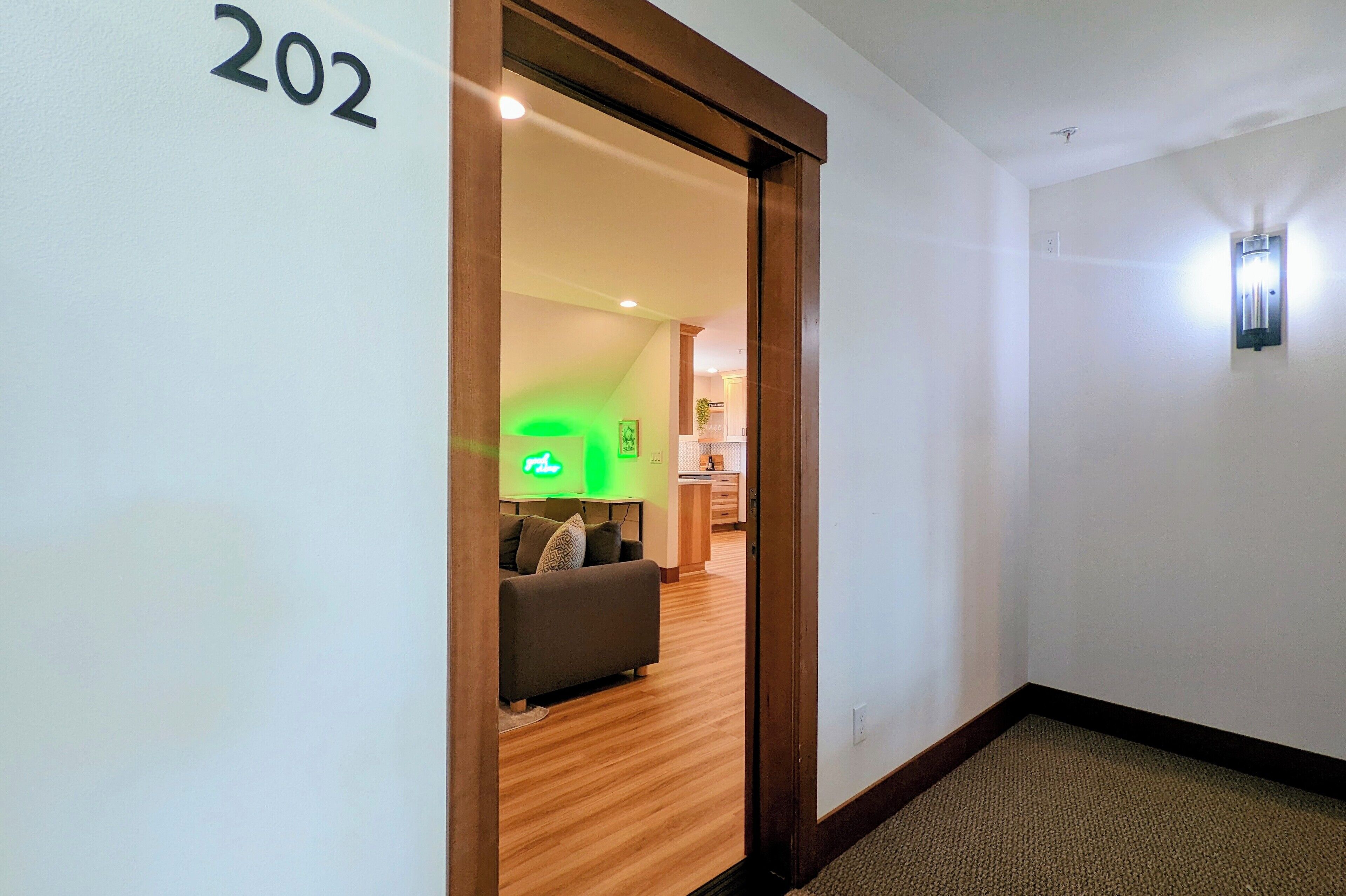 A hallway with a wooden door labeled "202" leading to a living area with a sofa and a neon sign.
