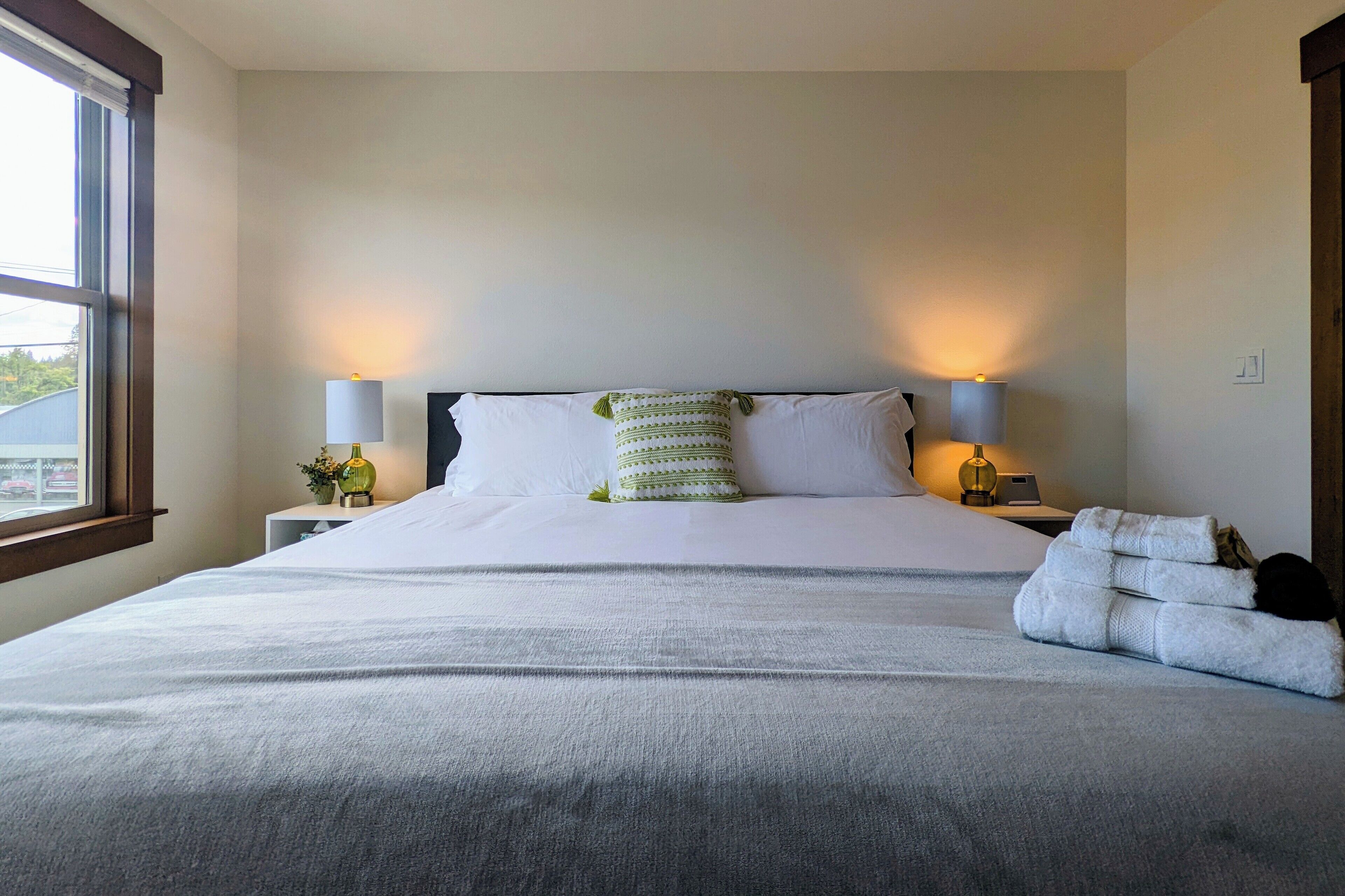 A neatly made bed with white linens, two bedside lamps, and a window with blinds.