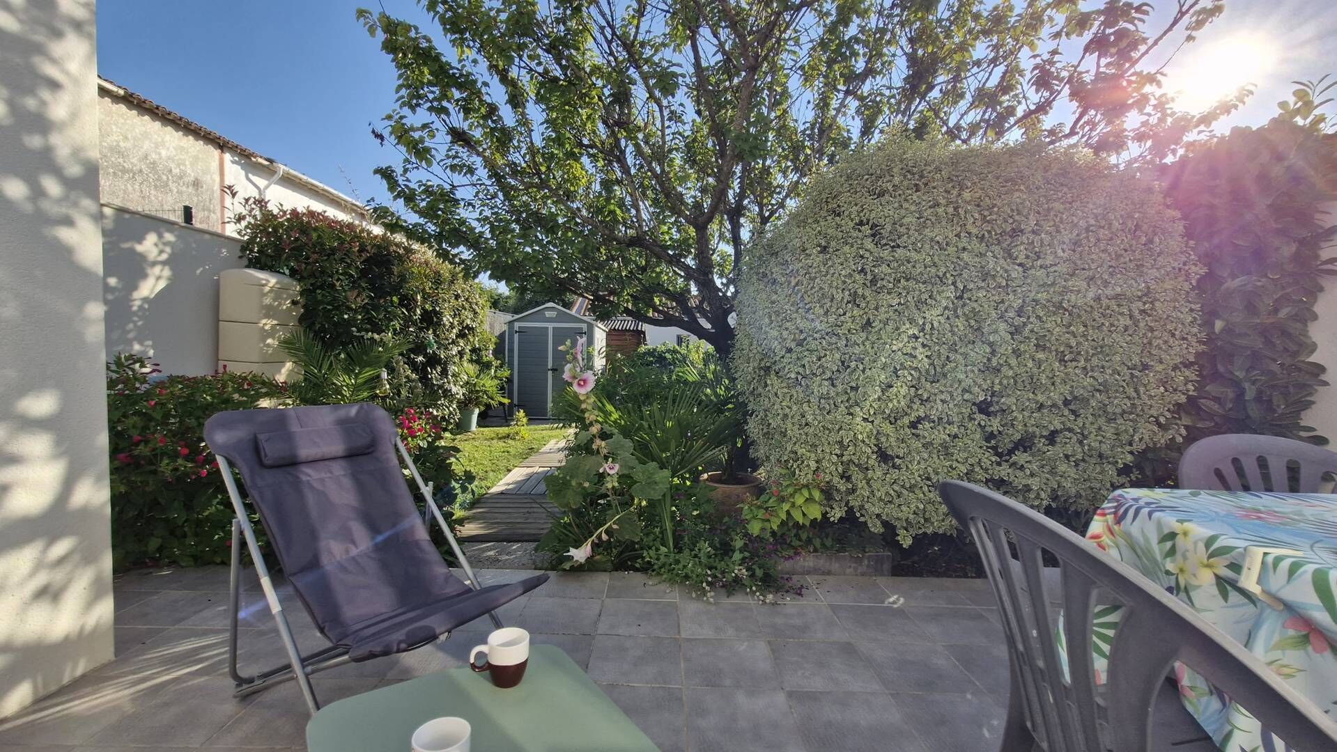 A patio with a folding chair, a table with two cups, a small garden, and a shed in the background.