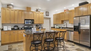 Una cocina moderna con gabinetes de madera, un refrigerador de acero inoxidable, un microondas y una estufa a gas. Hay tres taburetes de bar y una pequeña isla con un fregadero.