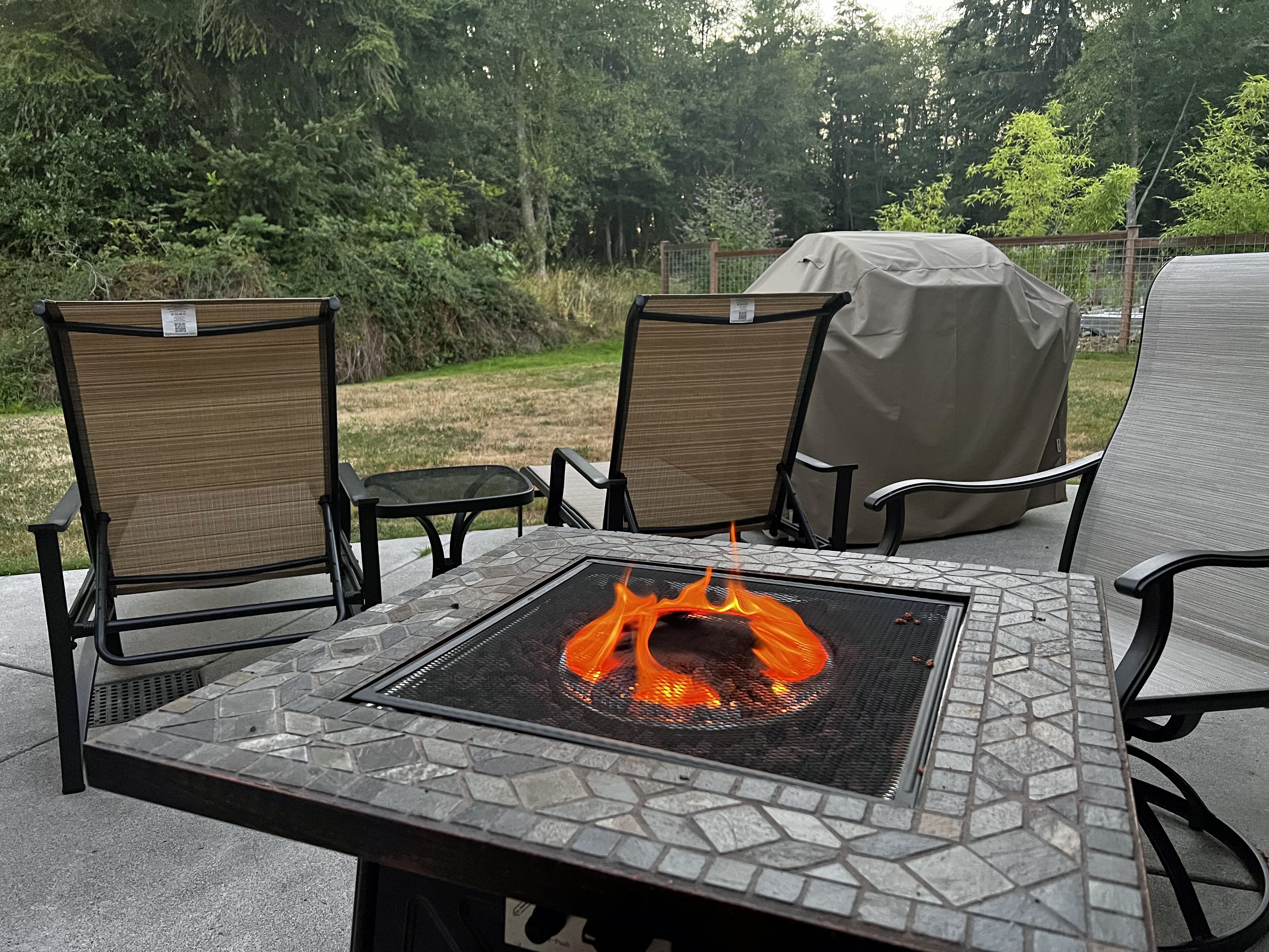 Een terras buiten met een vuurtafel van steen, twee comfortabele stoelen en een overdekte barbecue.