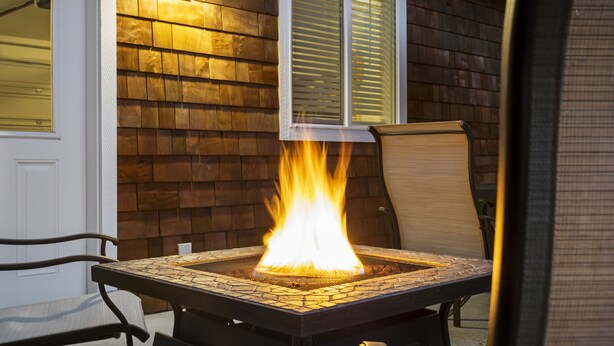 Una chimenea cuadrada al aire libre con un revestimiento de baldosas, ubicada frente a una pared de madera con una ventana y un paraguas rojo.