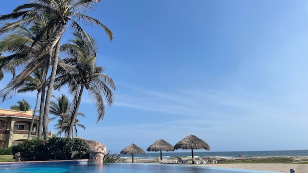 A resort pool with palm trees, thatched umbrellas, and a view of the ocean.