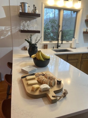A kitchen counter with a cheese board, a bowl of bananas, and a window with a view of trees.