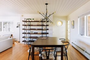 A dining area with a wooden table and chairs, a modern chandelier, and a bookshelf with decorative items.