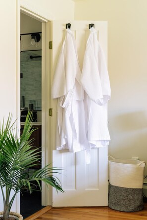 A white door with two white robes hanging on hooks, a potted plant, and a woven basket on the floor.