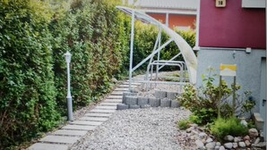 A pathway leading to a building with a red exterior, a metal staircase, and a small garden area with rocks and plants. - Apartment (Zislow)