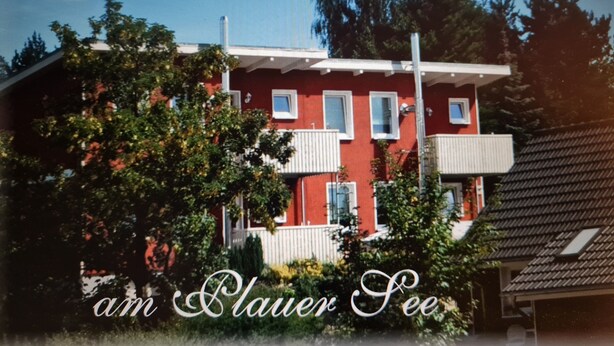 A red building with white windows and balconies, surrounded by trees and greenery. - Apartment (Zislow)
