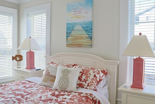 A bedroom with a bed, pillows, a nightstand, a lamp, a painting, and a window with blinds.
