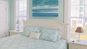 Ein Schlafzimmer mit einem Bett, einem Nachttisch, einer Lampe und einem Bild vom Meer.