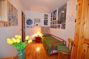 Une petite chambre à coucher avec un lit simple, une table de chevet, une lampe, une chaise et un vase de tulipes.