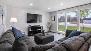 Una sala de estar moderna con un sofá seccional gris, un televisor de pantalla plana montado en la pared y un amplio ventanal con vistas al exterior.