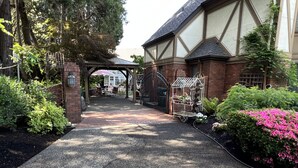 Ein Weg führt zu einem Gebäude mit überdachter Veranda, ein Metall-Gartencar mit Blumen steht daneben.