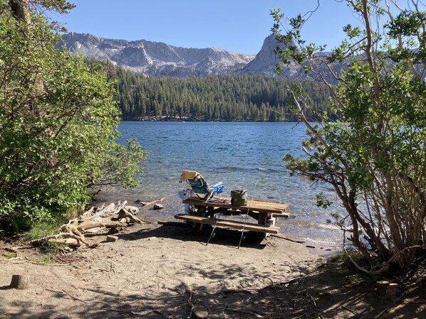 A lakeside picnic area with a bench and a cooler, surrounded by trees and mountains in the background.