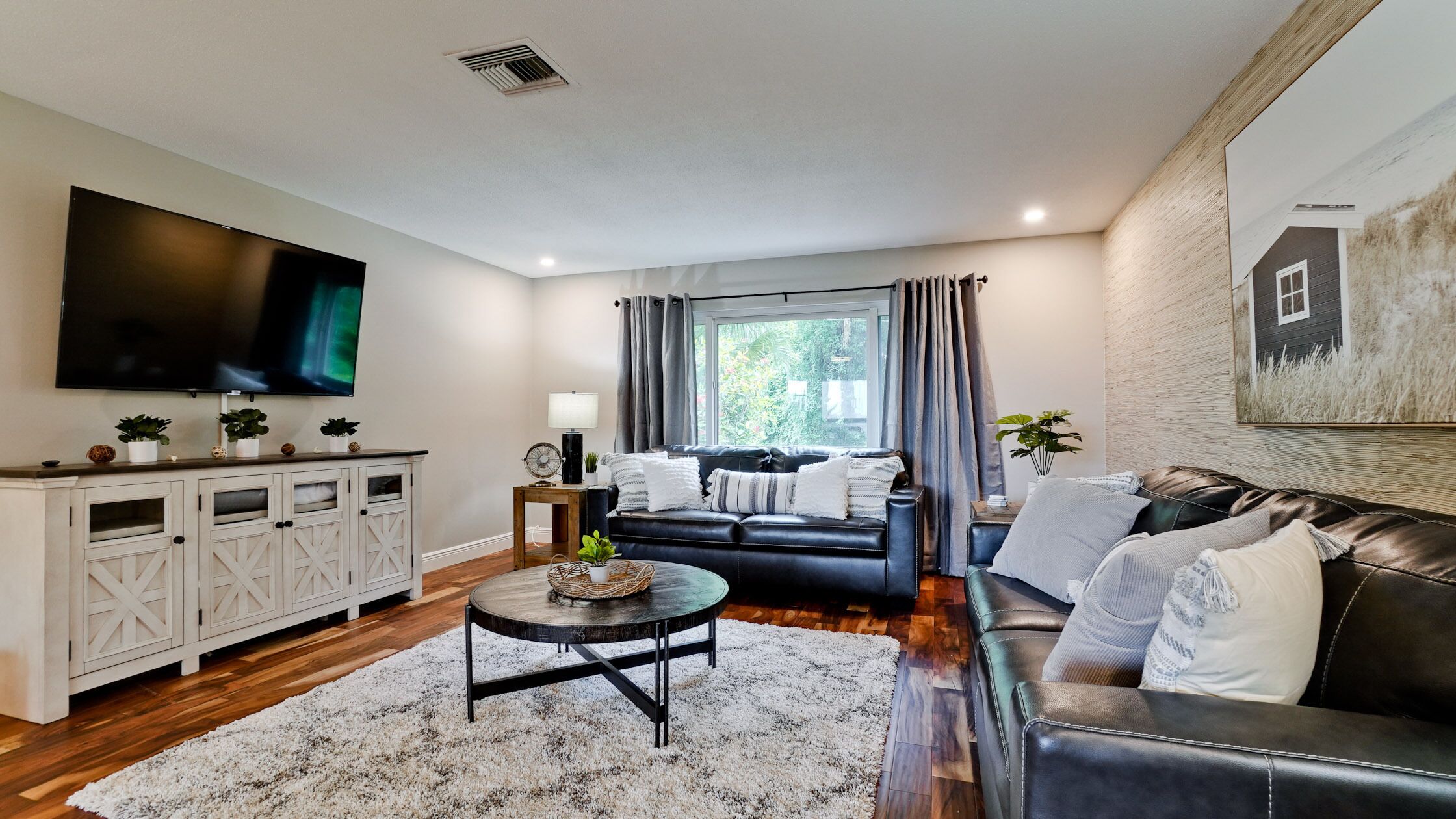 A modern living room with a black leather sofa, a white cabinet with glass doors, a flat-screen TV, and a large window with curtains.