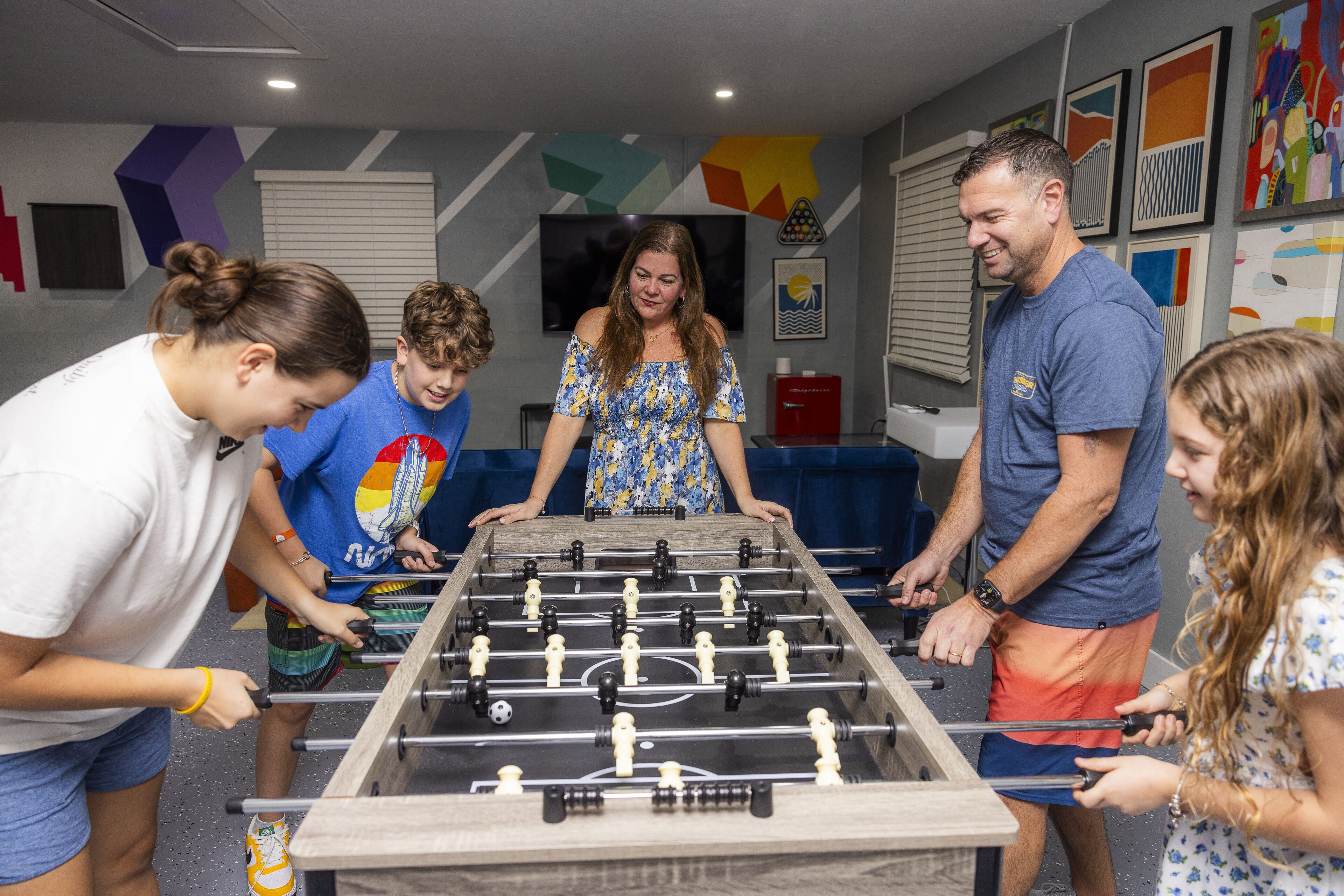 A group of people playing foosball in a room with a modern design, including geometric wall decorations and a flat-screen TV.