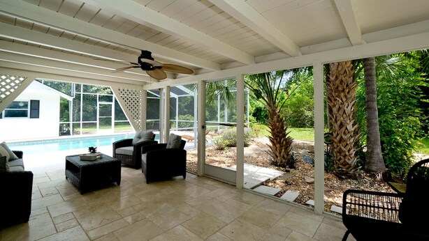 Ein überdachter Patio-Bereich mit Gartenmöbeln, einem Deckenventilator und Blick auf ein Schwimmbad und Garten.
